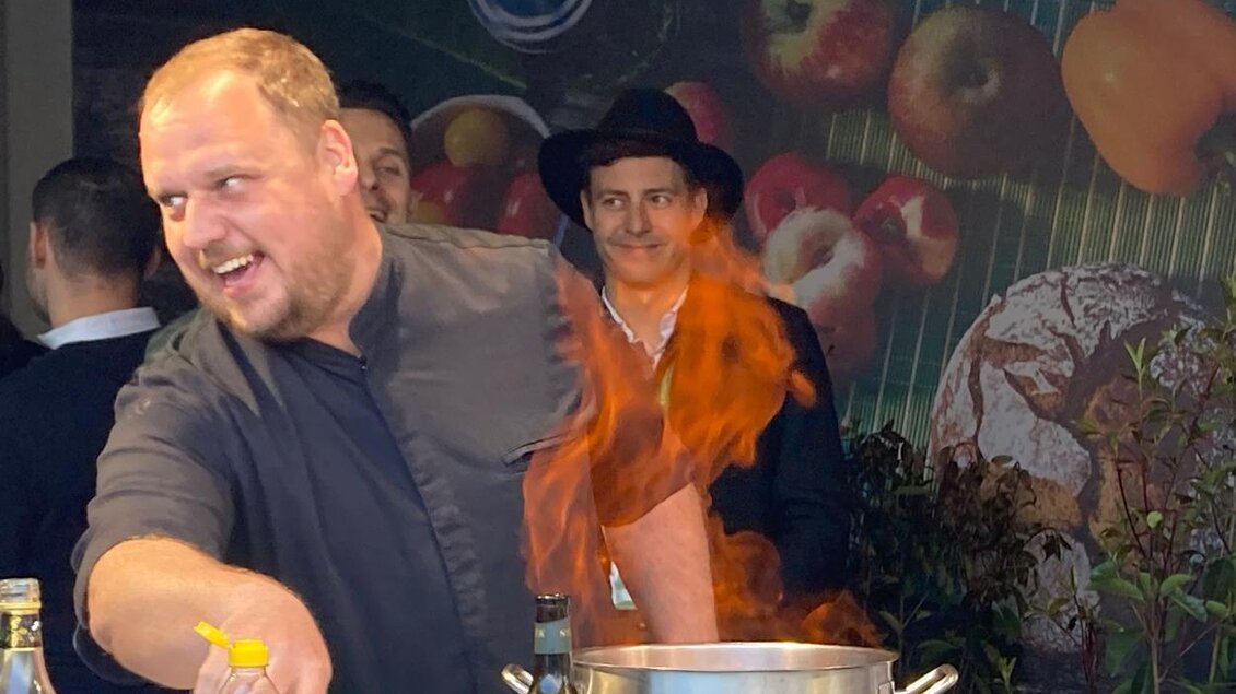 Ein Koch, der mit einem Lächeln in einer Pfanne flambiert, während er von einem Zuschauer im Hintergrund beobachtet wird. Im Hintergrund sind frische Äpfel und Gemüse zu sehen.