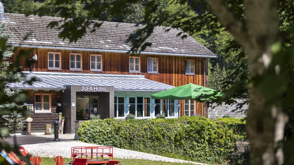 Ein gemütliches Holzhaus mit einem einladenden Eingangsbereich. Umgeben von grünen Büschen und Sonnenschirmen im Freien. | © Jufa Hotel Eisenerz