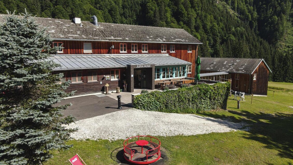 Ein rustikales Holzhaus mit einer modernen Terrasse inmitten grüner Landschaft. Rote Liegestühle und ein Feuerplatz laden zum Entspannen ein. | © Jufa Hotel Eisenerz