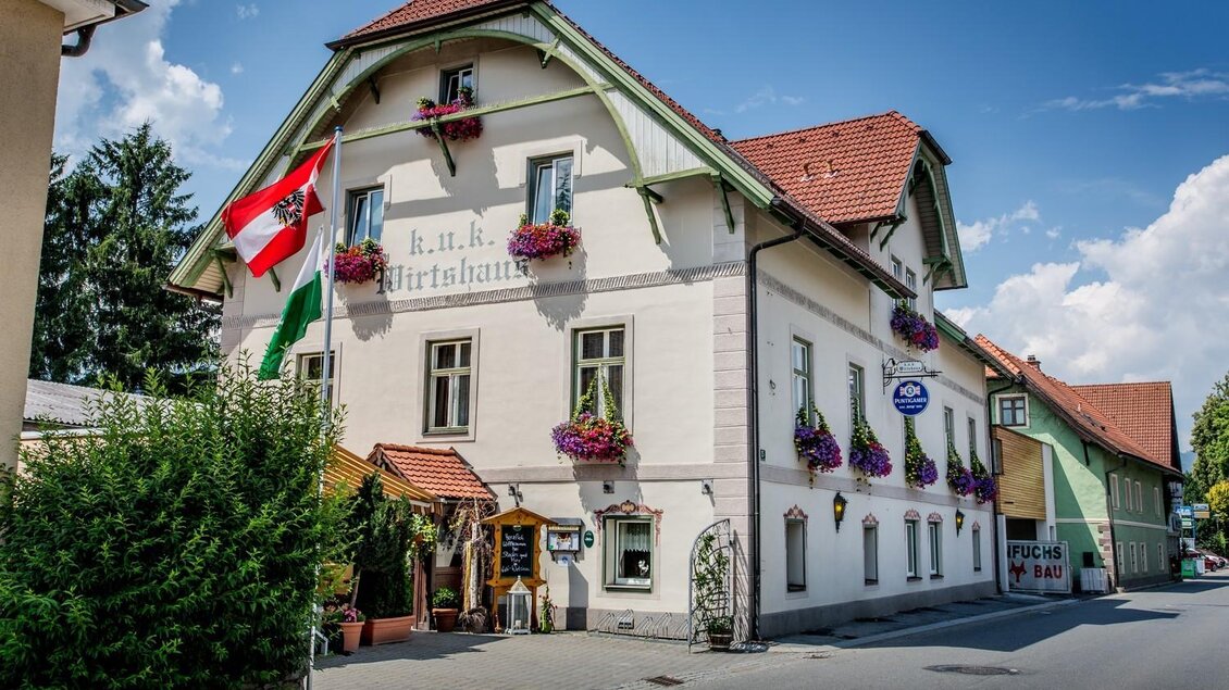 Ein hübsches Gasthaus mit bunten Blumen und Fahnen. Die Straßenansicht zeigt eine einladende Atmosphäre mit klar blauem Himmel. | © K.U.K. Wirtshaus