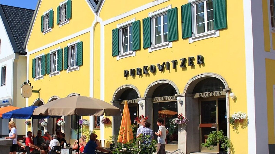 Schöne gelbe Gebäude im Stadtzentrum mit grünen Fensterläden. Draußen sitzen Menschen an Tischen und genießen die Sonne. | © Familie Purkowitzer