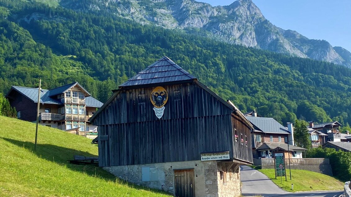 Ein traditionelles Holzhaus steht in einer malerischen Landschaft. Im Hintergrund erheben sich majestätische Berge und grüne Wiesen. | © TVB Ausseerland Salzkammergut/Bettina Scheck