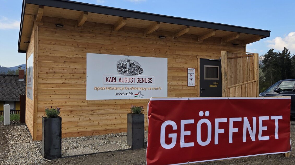 Ein Holzgebäude mit einem Schild, das "Karl August Genuss" zeigt. Vor dem Gebäude hängt ein großes Banner mit der Aufschrift "GEÖFFNET". | © Karl August
