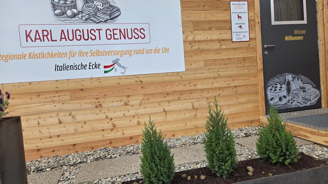 Ein Holzgebäude mit einem Schild, das „KARL AUGUST GENUSS“ und den Hinweis auf regionale Köstlichkeiten zeigt. Der Eingangsbereich ist mit kleinen Pflanzen und einem freundlichen Willkommen-Schild dekoriert. | © Karl August