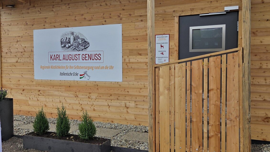 Ein Holzgebäude mit einem modernen Eingang und einem Schild mit der Aufschrift "KARL AUGUST GENUSS". Vor dem Eingang stehen einige kleine Pflanzen in einem Pflanzkasten. | © Karl August