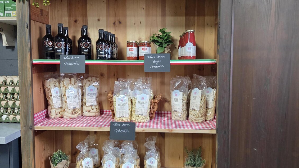 Ein Holzregal mit verschiedenen Produkten, darunter Öle, Pasta und Dekoration. Die Regale sind dekorativ mit grünen Pflanzen und Beschriftungen gestaltet. | © Karl August