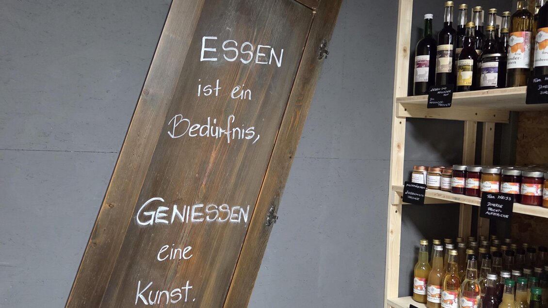 Eine Holztafel mit der Aufschrift „ESSEN ist ein Bedürfnis, GENIESSEN eine Kunst“ hängt an der Wand. Im Hintergrund sind Regale mit verschiedenen Gläsern und Flaschen zu sehen. | © Karl August