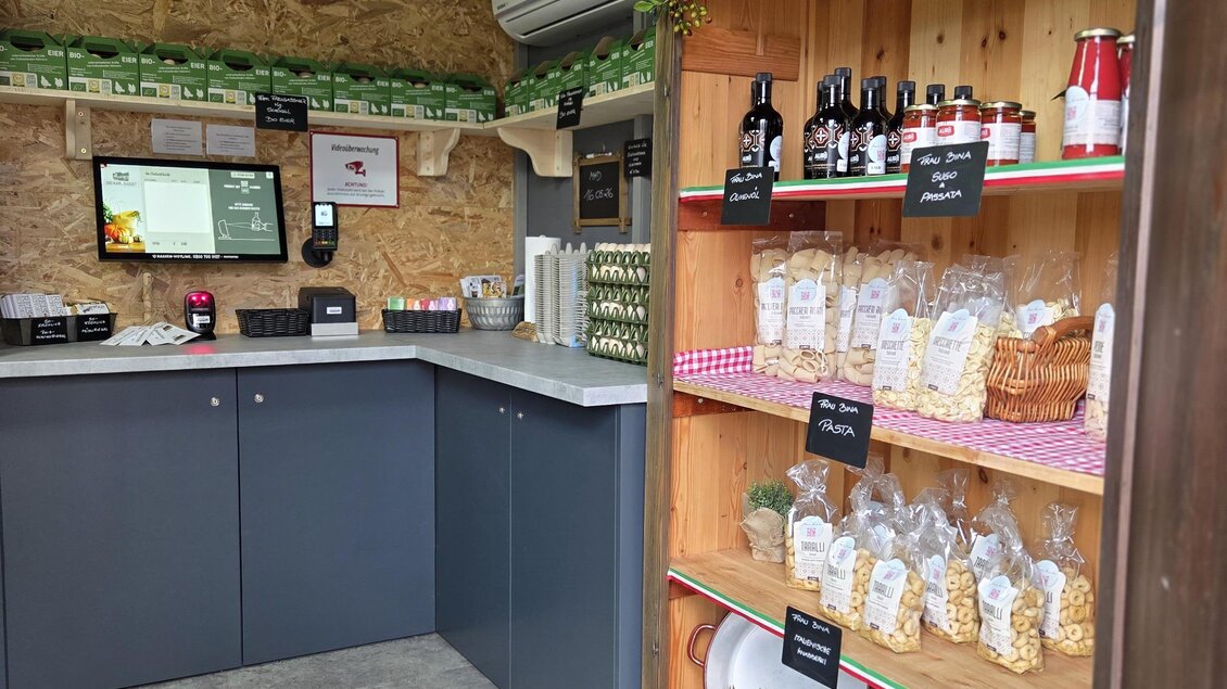 Ein kleines Geschäft mit einem Holzregal voller Produkte wie Wein und Nudeln. Die Wand ist mit grünen Zweigen dekoriert und es gibt einen modernen Tresen. | © Karl August