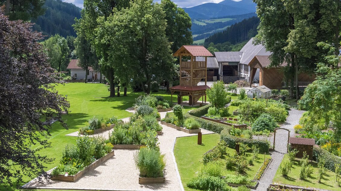 Ein schöner Garten mit verschiedenen Pflanzen und Wegen. Im Hintergrund sind Bäume und ein Gebäude zu sehen, umgeben von einer malerischen Landschaft. | © Anita Fössl