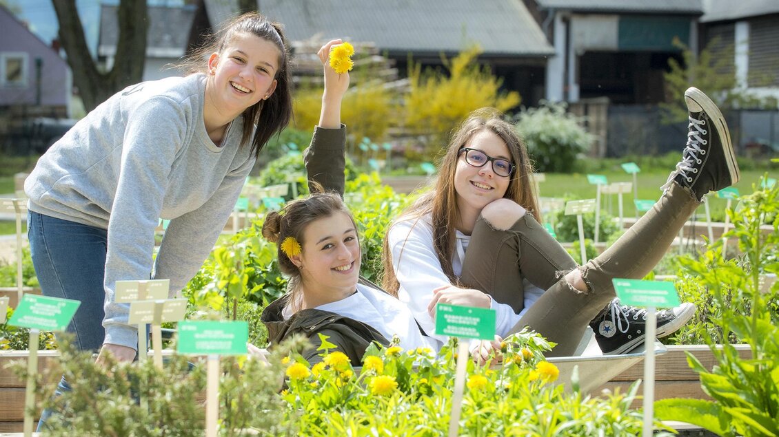 Drei junge Frauen lächeln fröhlich in einem Garten zwischen bunten Pflanzen. Sie sitzen und stehen inmitten von Hochbeeten und genießen die sonnige Atmosphäre. | © Anita Fössl