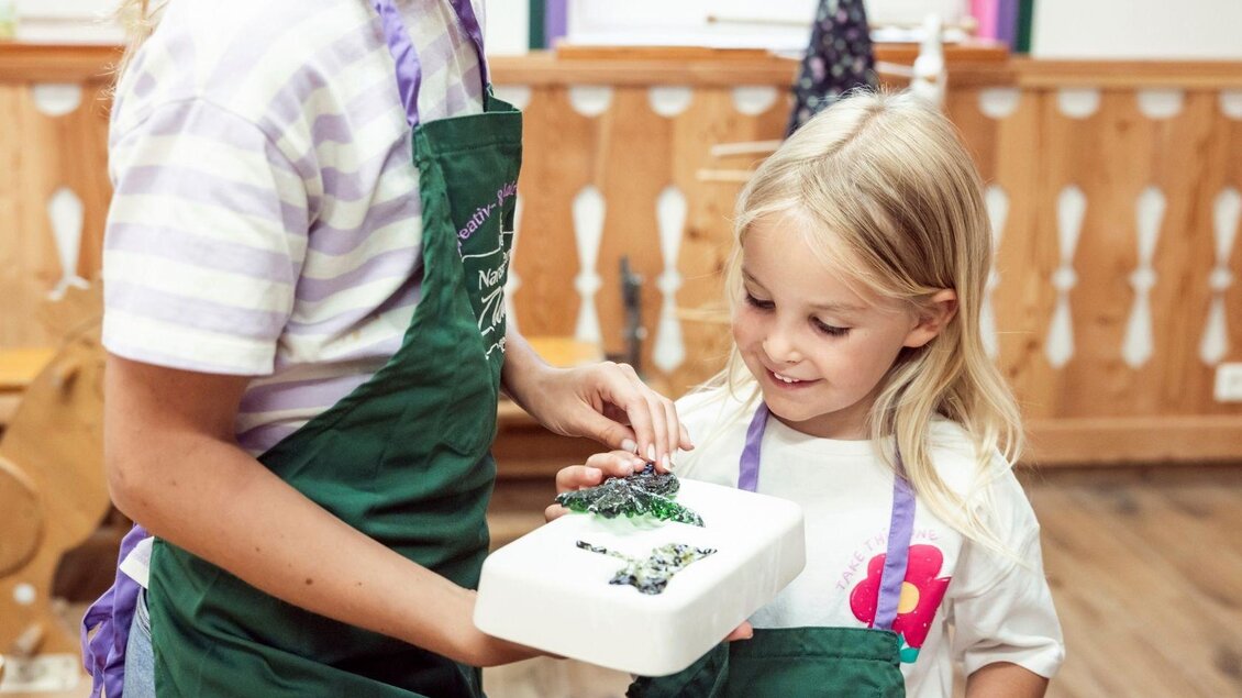 Ein Mädchen und eine Betreuerin zeigen gemeinsam eine Torte. Beide tragen grüne Schürzen und lächeln. | © www.zloam.at
