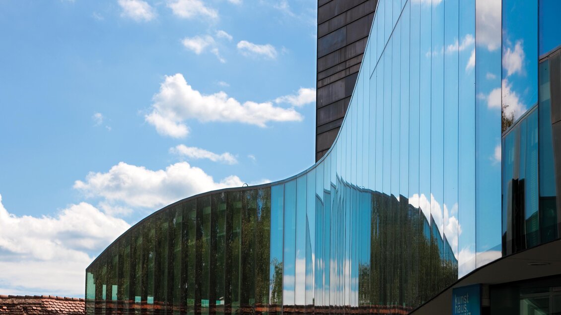 Ein modernes Gebäude mit gläserner Fassade, das den Himmel und die Wolken reflektiert. Die geschwungene Form verleiht dem Architekturdesign einen dynamischen Eindruck. | © Kunsthaus Weiz