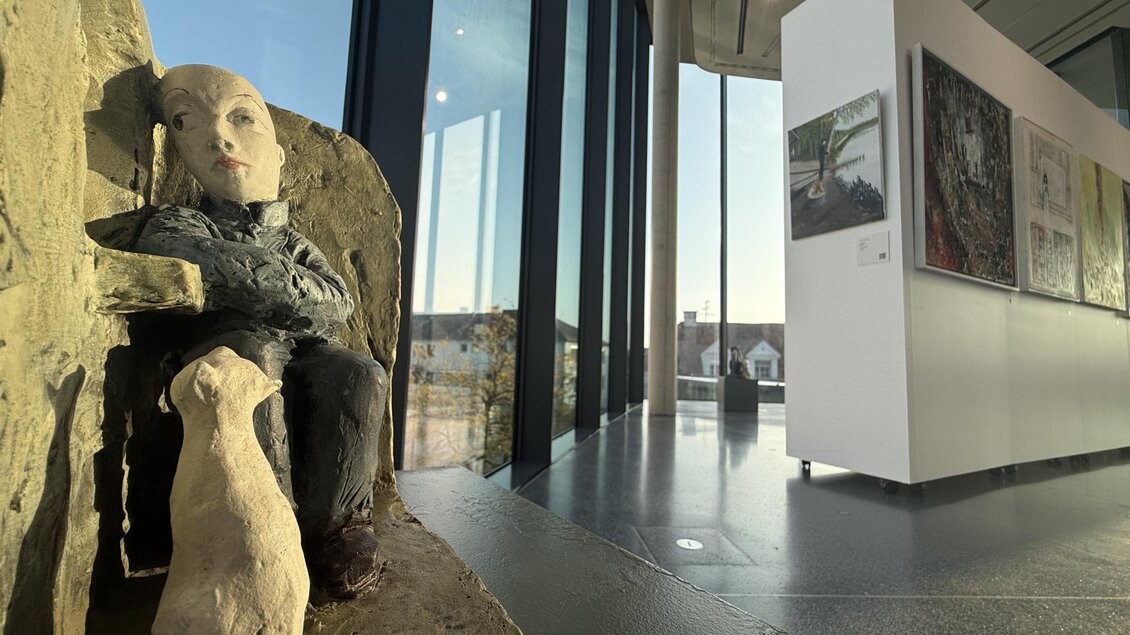 Eine Skulptur eines sitzenden Menschen mit einem Hund in einem modernen Ausstellungsraum. Im Hintergrund sind große Fenster und mehrere Bilder an der Wand zu sehen. | © Kunsthaus Weiz