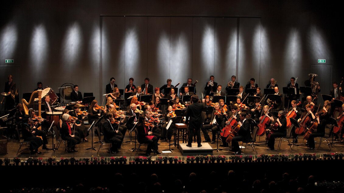 Ein großes Orchester spielt auf einer Bühne. Der Dirigent führt die Musiker mit Begeisterung. | © Kunsthaus Weiz