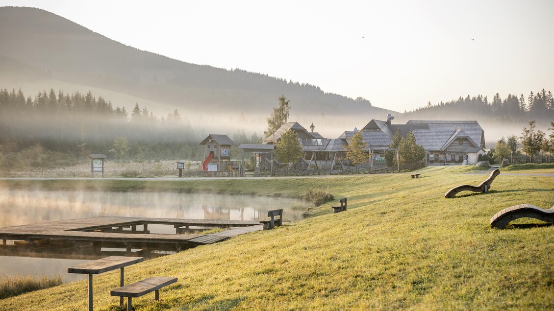 Eine ruhige Landschaft mit einem Teich und einem Nebel, der die Umgebung umhüllt. Im Hintergrund sind Berge und ein charmantes Gebäude mit Bäumen zu sehen. | © Latschenhütte