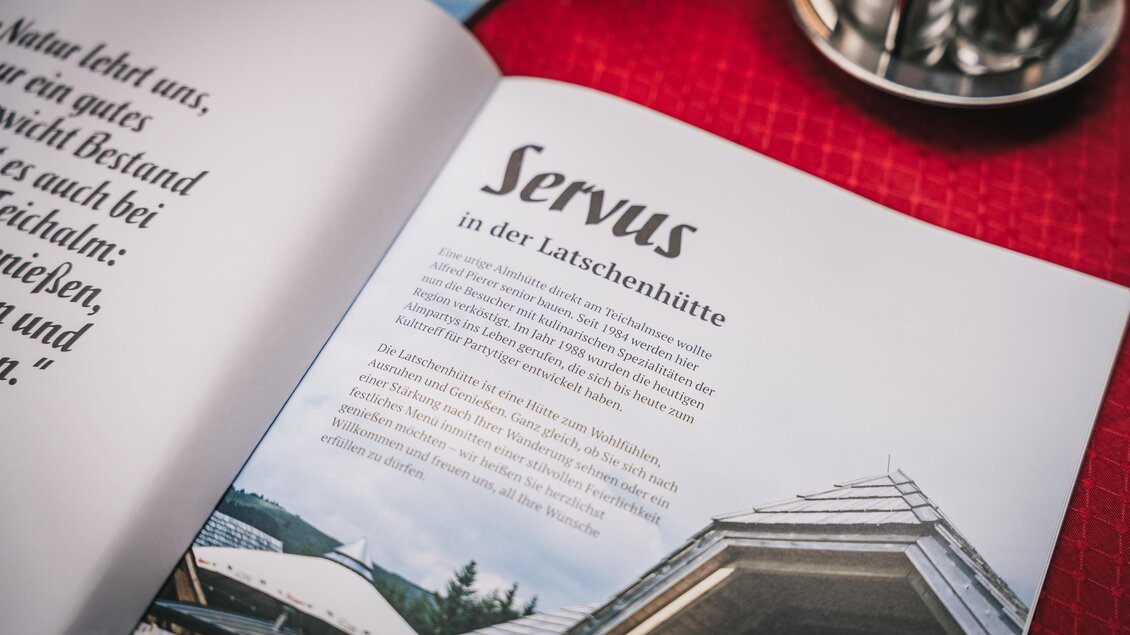 Ein offenes Buch mit einer Seite, die das Wort "Servus" zeigt. Im Hintergrund ist eine Holzarchitektur zu sehen, die zu einer Hütte gehört. | © Oststeiermark Tourismus