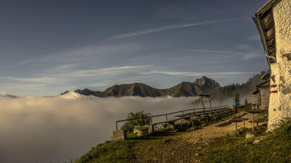 Eine atemberaubende Berglandschaft über einer Wolkendecke, mit beeindruckenden Gipfeln im Hintergrund. Im Vordergrund sieht man eine Terrasse mit Sitzgelegenheiten und einen Blick auf die umliegende Natur. | © Leobner Hütte