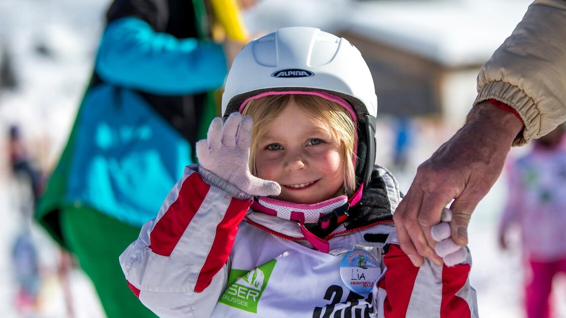 <p>Ein lächelndes kleines Mädchen in Skikleidung winkt fröhlich in die Kamera. Im Hintergrund sind weitere Skifahrer am Loser in Altaussee zu sehen.</p> | © TVB Ausseerland - Salzkammergut/T. Lamm