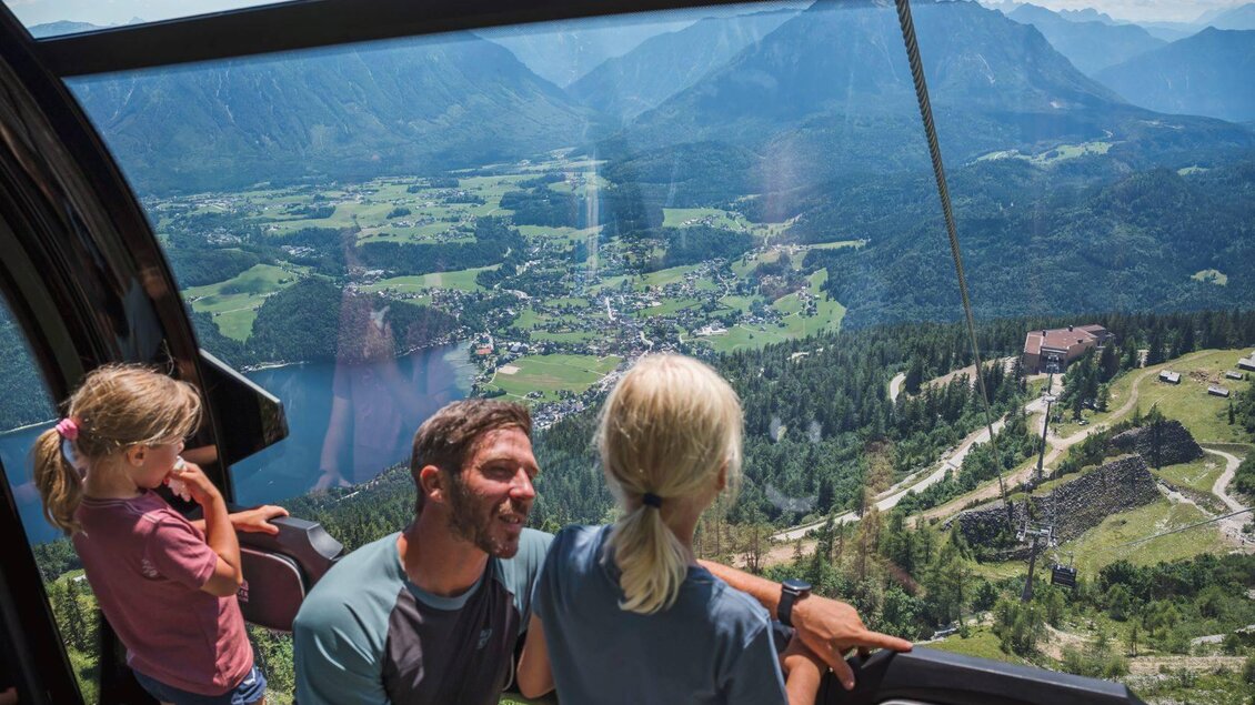 Eine Familie fährt in einer Gondel mit Blick auf eine malerische Landschaft. Im Hintergrund sind Berge, Wälder und ein Dorf am See zu sehen. | © Klaus Krumböck