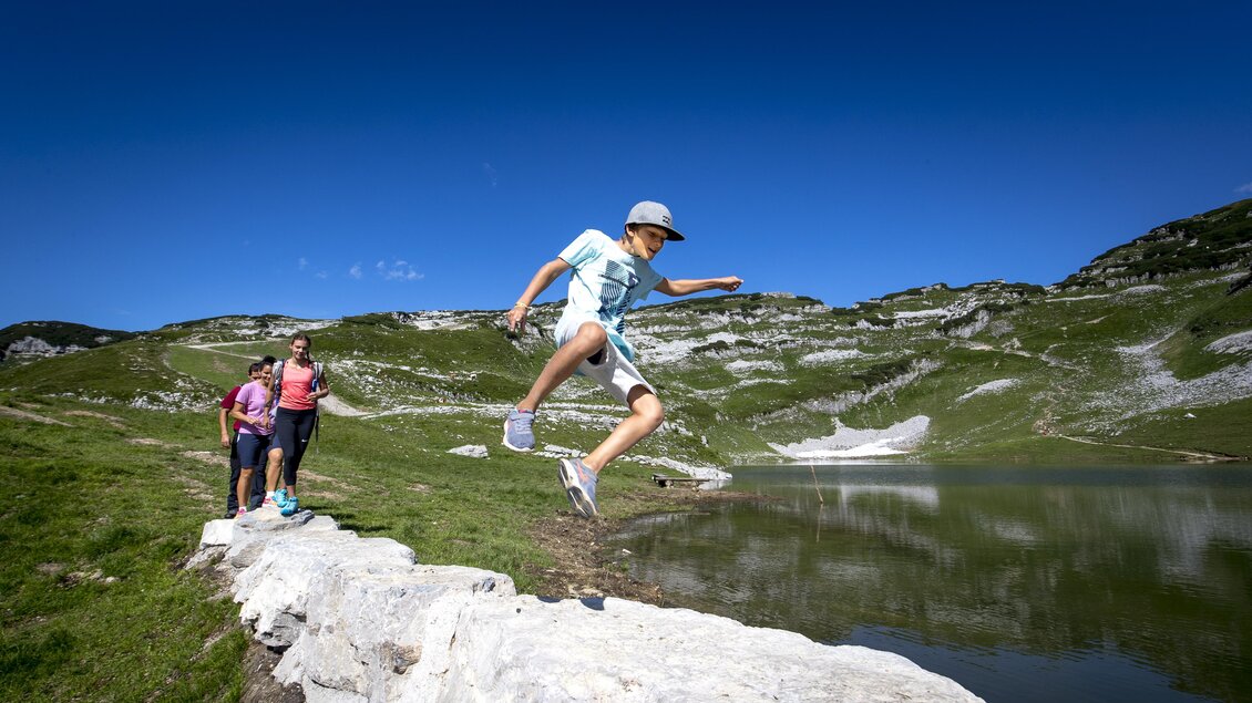 <p>Ein Junge springt fröhlich über einen Stein beim malerischen Augstsee am Loser in Altaussee. Im Hintergrund sind Wanderer unterwegs, welche durch die schöne sommerliche Berglandschaft des Hausberges der Altausseer spazieren.</p> | © TVB Ausseerland-Salzkammergut/Tom Lamm