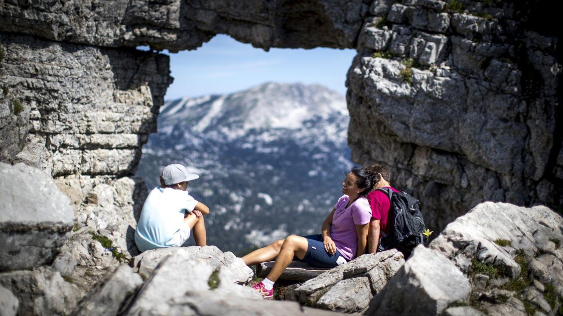<p>Drei Personen sitzen am Loserfenster in Altaussee und genießen die Aussicht durch den natürlichen Felsbogen am Loser. Die sommerliche Berglandschaft ist beeindruckend und die Stimmung entspannt.</p> | © TVB Ausseerland - Salzkammergut-Tom Lamm