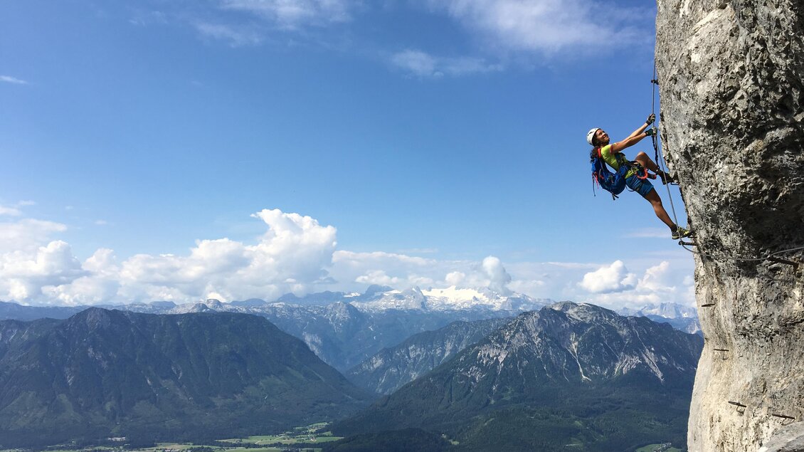 <p>Ein Kletterer klettert eine Felswand beim Loser-Panorama-Klettersteig Sisi hoch. Im Hintergrund erstreckt sich die beeindruckende Landschaft des Ausseerlandes mit dem Dachstein in der Ferne und einem blauen Himmel.</p> | © Loser Bergbahnen Altaussee/M. Raich