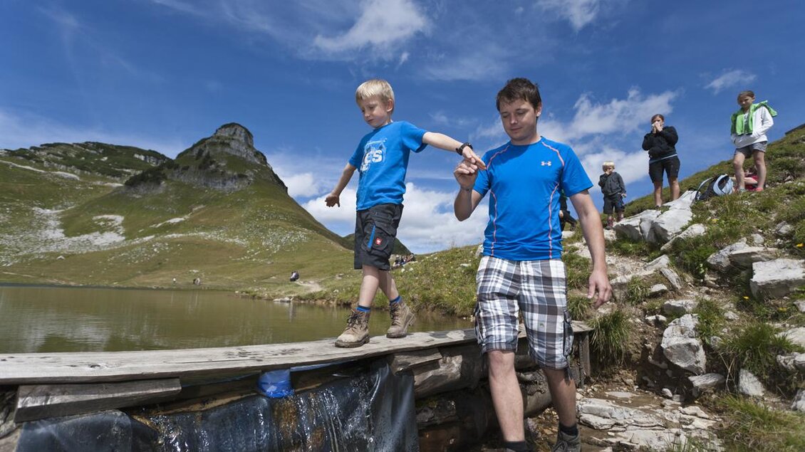 <p>Ein Mann hilft einem Jungen über ein Holzbrett am Rande des Augstsees am Loser zu balancieren. Im Hintergrund sind weitere Personen und die beeindruckende Berglandschaft des Ausseerlandes mit dem Atterkogel zu sehen.</p> | © Loser Bergbahnen Altaussee/Leo Himsl