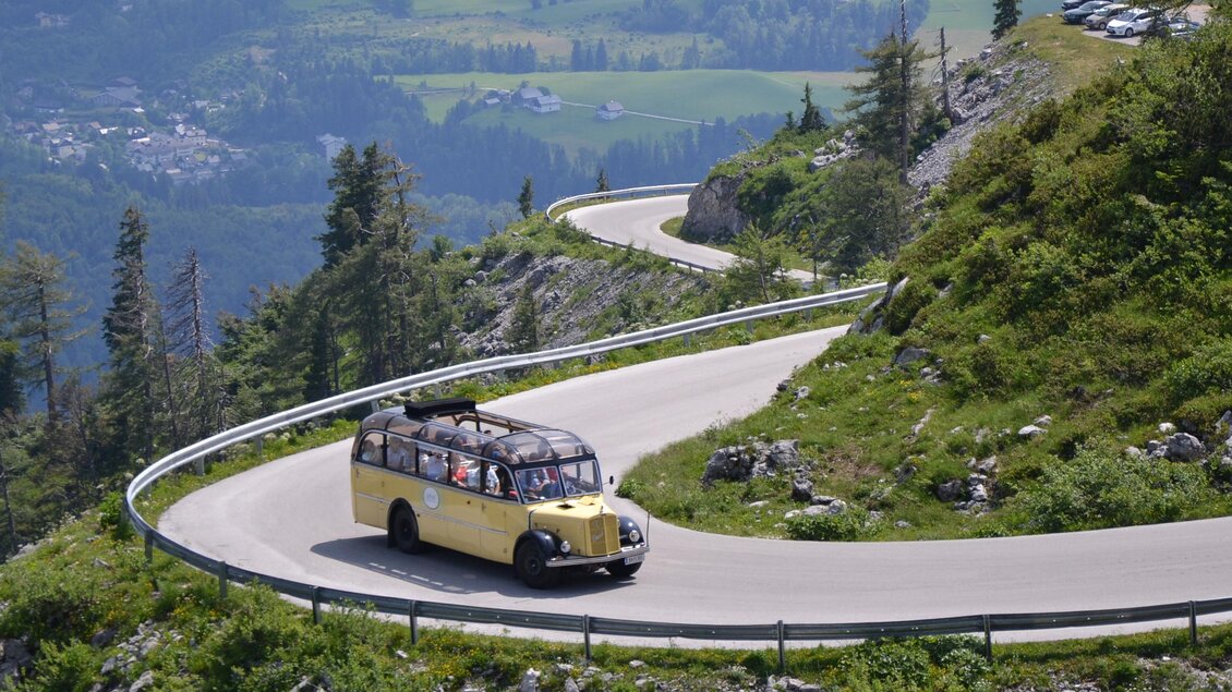 Ein gelbes Fahrzeug fährt eine kurvenreiche Straße inmitten von grünen Hügeln. Im Hintergrund sind Berge und die Aussicht auf das Tal sichtbar. | © Loser Bergbahnen