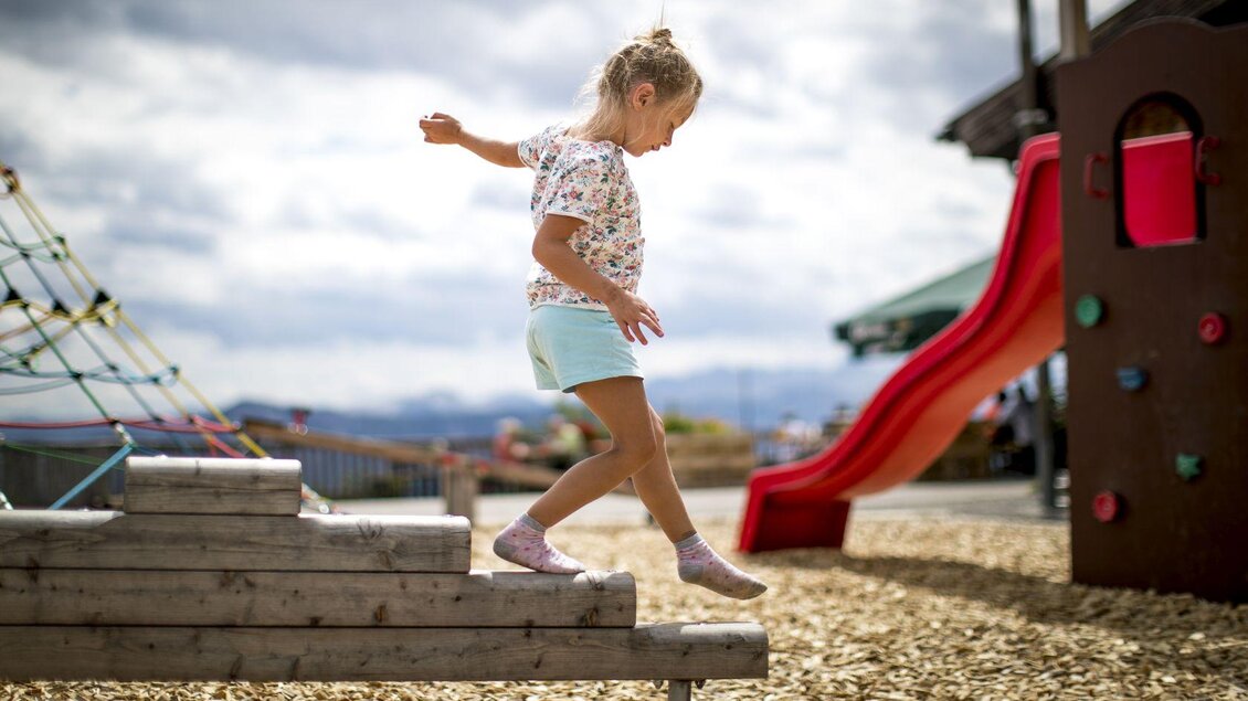 Ein Mädchen balanciert auf einem Holzelement auf dem Spielplatz. Im Hintergrund ist eine Rutsche und eine Kletterstruktur zu sehen. | © TVB Ausseerland - Salzkammergut-Tom Lamm