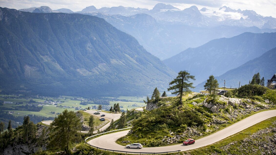 Eine kurvenreiche Straße führt durch eine malerische Berglandschaft. Im Hintergrund erheben sich majestätische Berge unter einem bewölkten Himmel. | © TVB Ausseerland - Salzkammergut-Tom Lamm