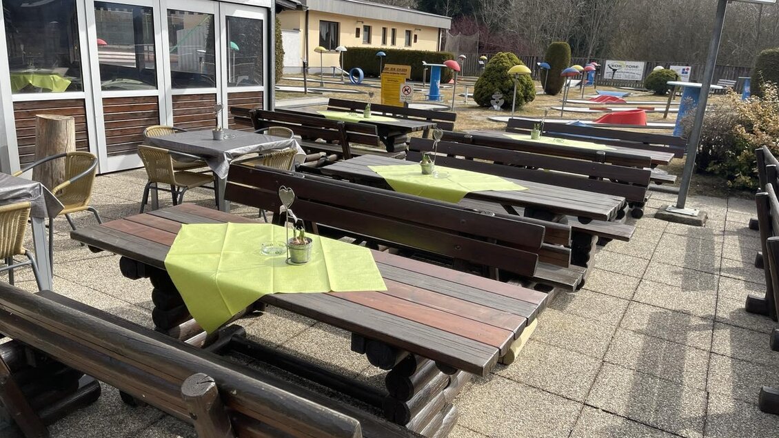 Eine Terrasse mit Holzbank-Tischen und grünen Tischdecken. Im Hintergrund sind Gebäude und gepflegte Sträucher zu sehen. | © Landhotel Reitingblick