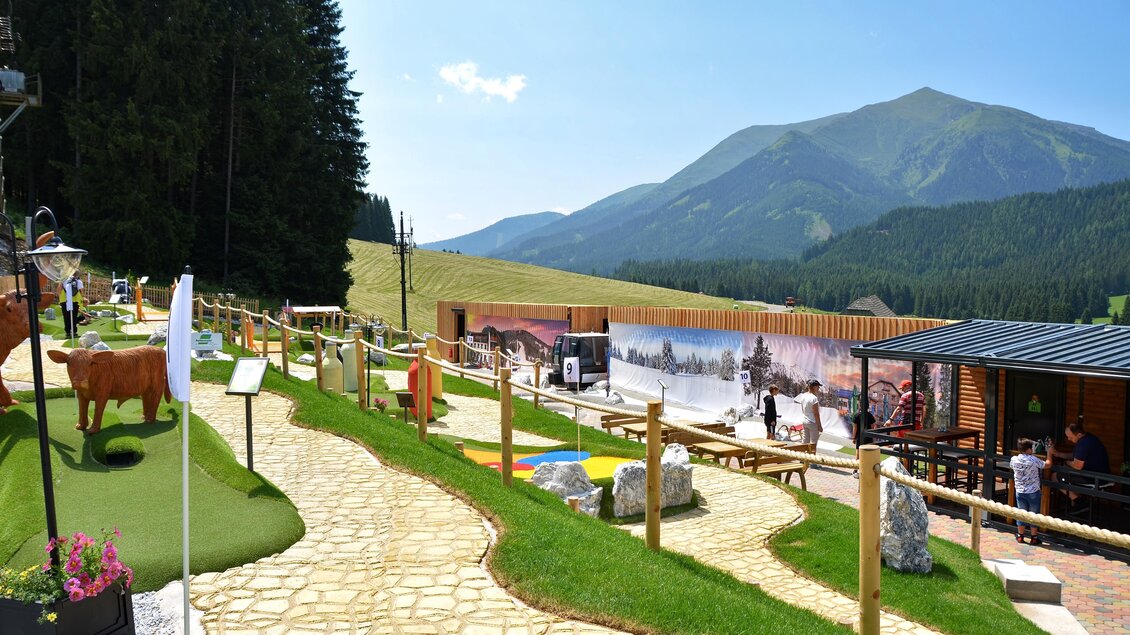 Eine malerische Landschaft mit einem Wanderweg, umgeben von Bäumen und Bergen. Im Vordergrund sind einige Spielmöglichkeiten und ein rustikales Gebäude zu sehen. | © Mountain Adventure Golf