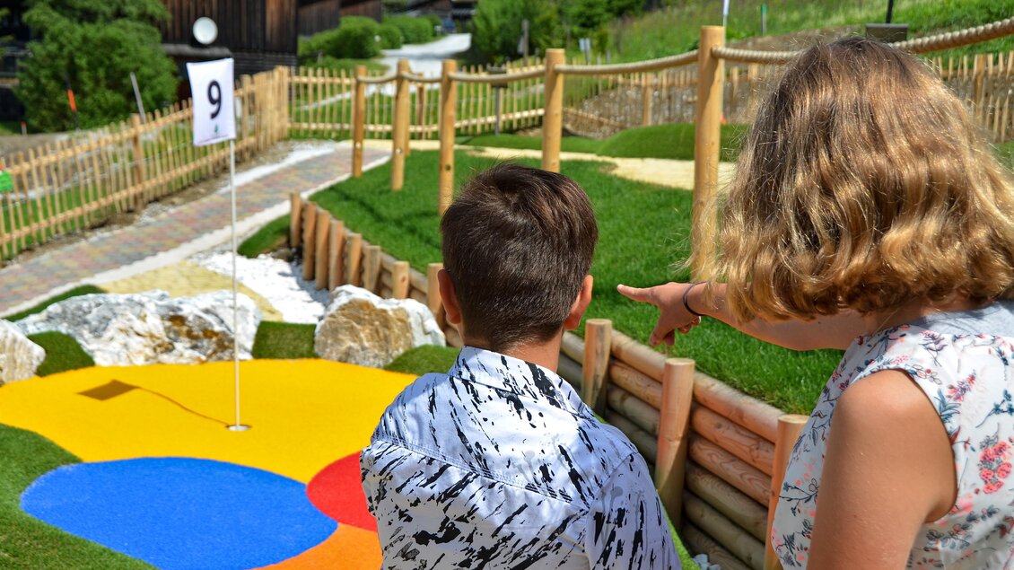 Kinder spielen Minigolf auf bunten Bahnen. Im Hintergrund sind grüne Wiesen und ein Holzzaun zu sehen. | © Mountain Adventure Golf