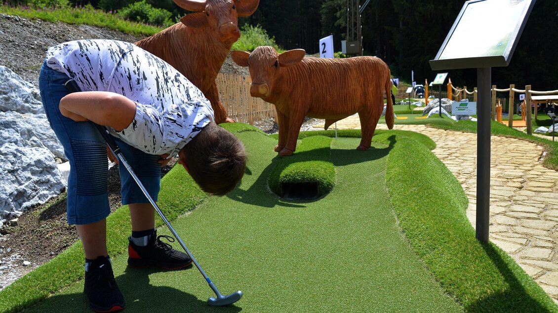 Ein Junge spielt Minigolf auf einem grünen Platz. Neben ihm stehen zwei Tierfiguren, die wie Highland-Rinder aussehen. | © Mountain Adventure Golf