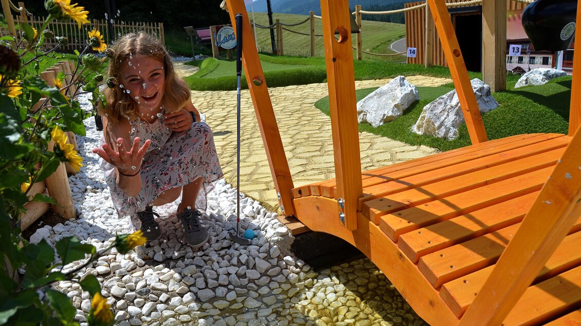 Ein Mädchen sitzt neben einem Holzsteg und lächelt. Es gibt Blumen und Steine um sie herum, sowie eine grüne Landschaft im Hintergrund. | © Mountain Adventure Golf