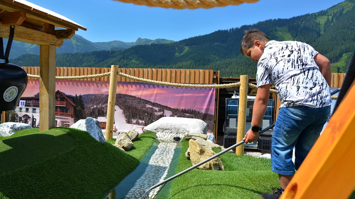Ein Junge spielt Minigolf in einer schönen Berglandschaft. Im Hintergrund sind grüne Hügel und ein blauer Himmel zu sehen. | © Mountain Adventure Golf