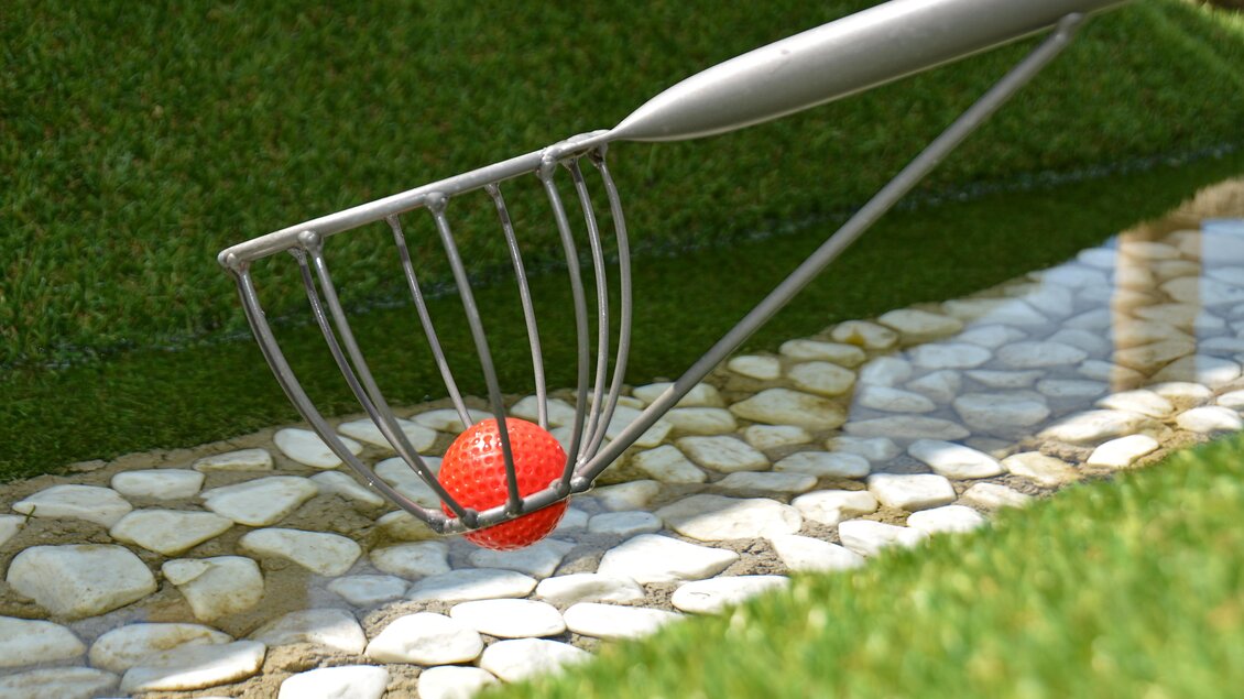 Ein Rasenrechen hebt einen roten Ball aus einem kleinen Gewässer. Umgeben von Kiessteinen und grünem Gras. | © Mountain Adventure Golf