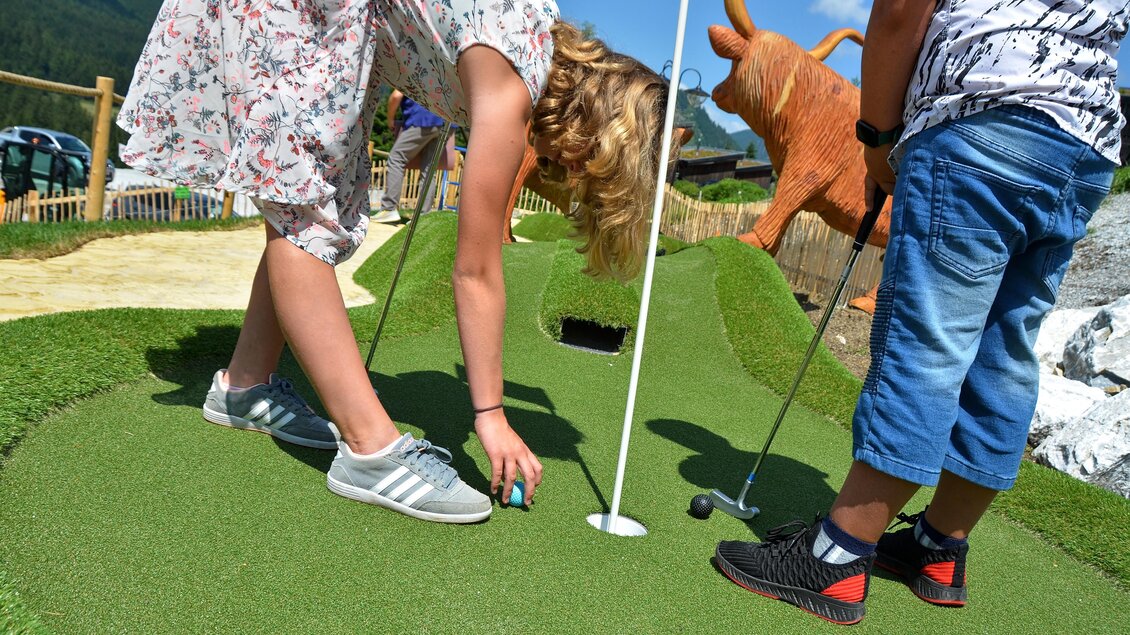 Ein Minigolfplatz im Freien mit zwei Kindern, die gerade spielen. Im Hintergrund sind dekorative Figuren und grüne Rasenflächen zu sehen. | © Mountain Adventure Golf