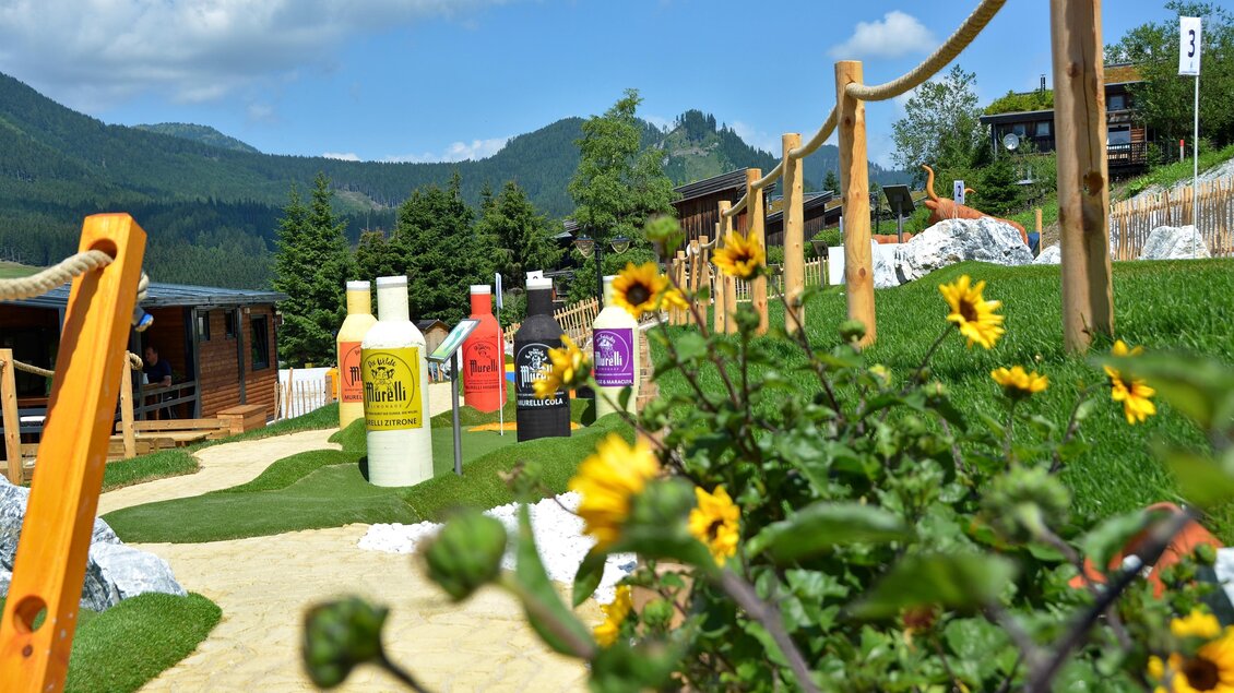 Ein malerischer Gartenweg mit bunten Flaschen und blühenden Sonnenblumen. Im Hintergrund sind sanfte Berge und ein klarer Himmel zu sehen. | © Mountain Adventure Golf