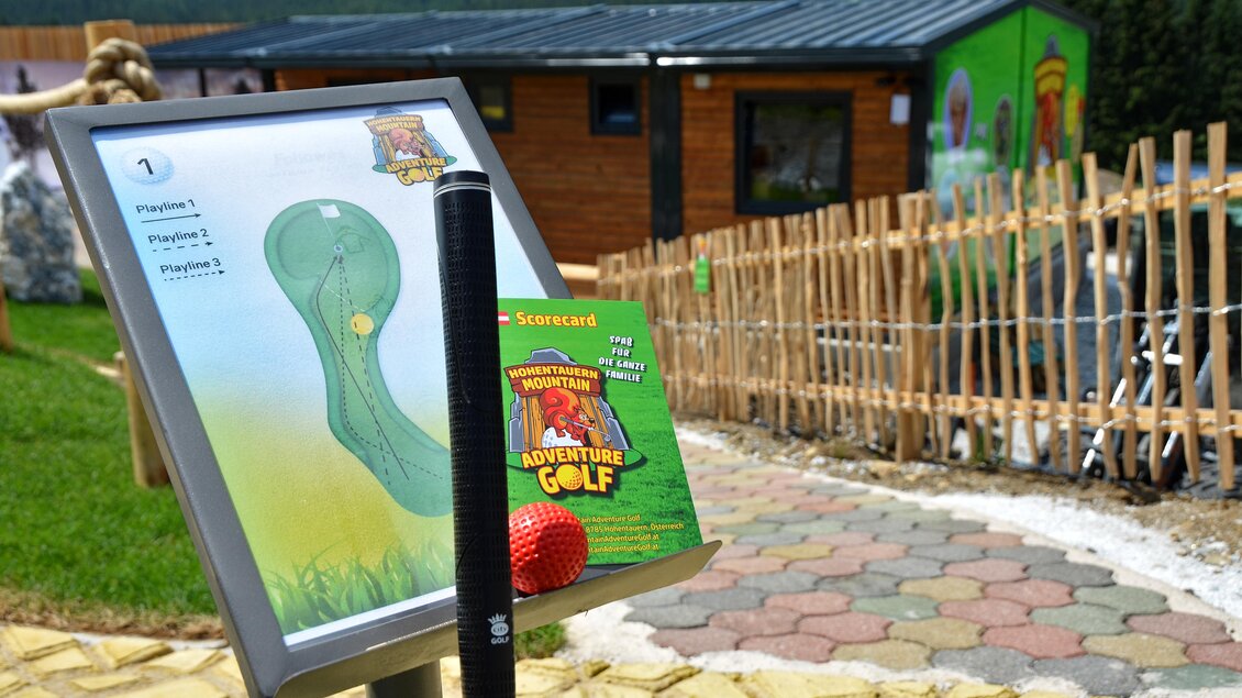 Ein Info-Schild zeigt die Bahn für Adventure Golf an. Im Vordergrund sind ein Golfball und ein Golfschläger sichtbar, während im Hintergrund ein Holzzaun und ein Gebäude zu erkennen sind. | © Mountain Adventure Golf