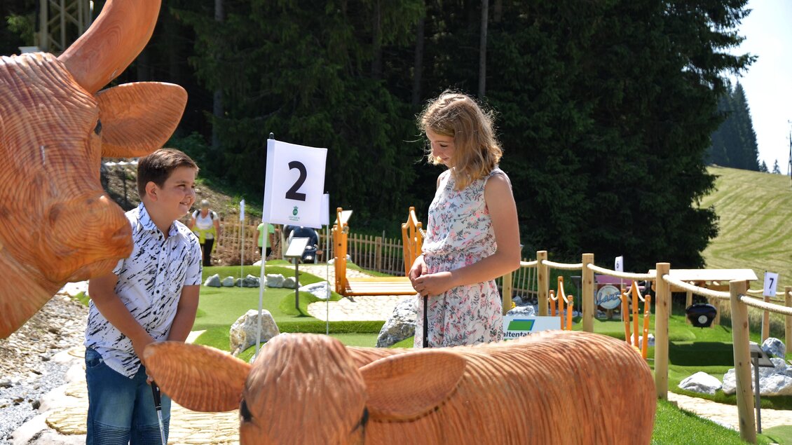 Zwei Kinder stehen neben großen Holzskulpturen von Kühen auf einem Minigolfplatz. Im Hintergrund sind grüne Wiesen und eine klare Atmosphäre zu sehen. | © Mountain Adventure Golf