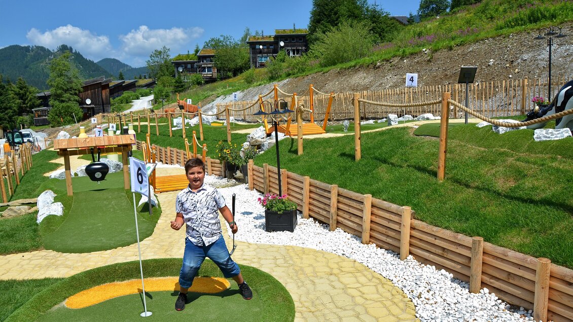 Ein Junge steht fröhlich auf einem Minigolfplatz mit verschiedenen Bahnen. Die grüne Landschaft ist von Bäumen und Hügeln umgeben. | © Mountain Adventure Golf