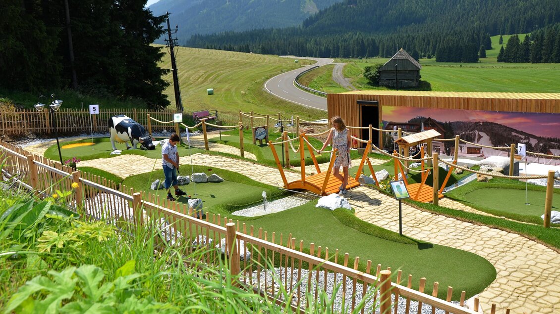 Eine Minigolfanlage in einer malerischen Berglandschaft. Im Vordergrund sind Spielgeräte und eine gedeckte Landschaft mit einer Kühe zu sehen. | © Mountain Adventure Golf