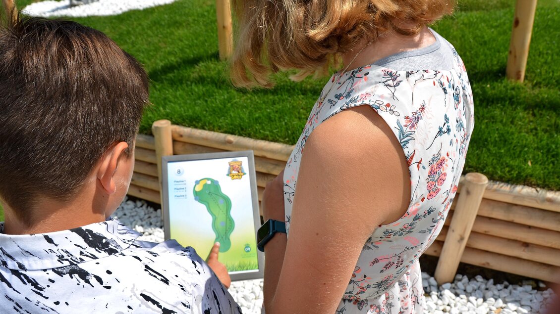 Ein Junge und ein Mädchen schauen auf ein Tablet. Im Hintergrund sieht man grünes Gras und einige Steine. | © Mountain Adventure Golf