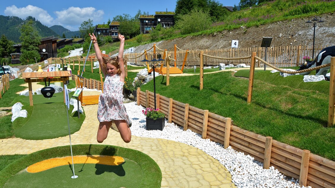 Ein Minigolfplatz mit grünen Rasenflächen und Holzbarrieren. Ein Mädchen springt fröhlich in der Luft. | © Mountain Adventure Golf
