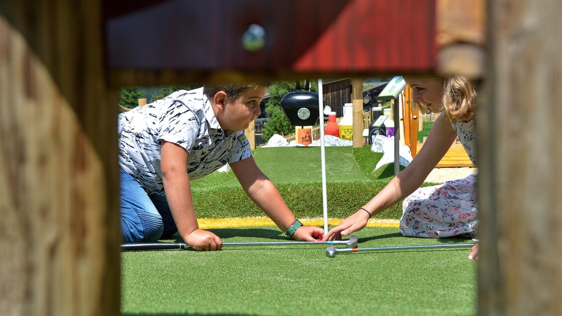 Zwei Kinder spielen Minigolf auf einem grünen Platz. Sie konzentrieren sich auf den Ball und die Fahne. | © Mountain Adventure Golf