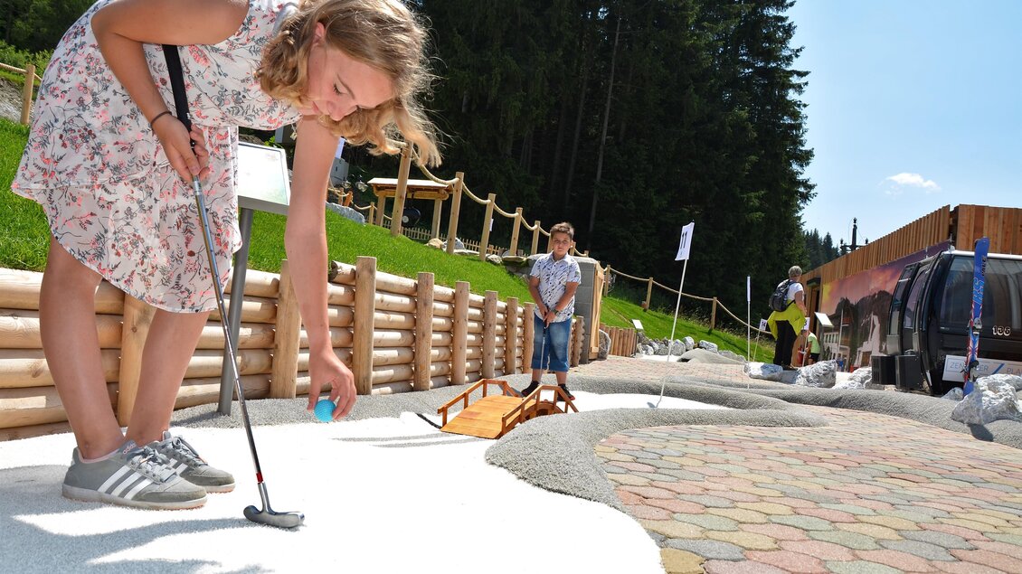 Ein Mädchen spielt Mini-Golf und zielt sorgfältig auf den Ball. Im Hintergrund ist ein Junge zu sehen, der ebenfalls an einer Spielaktivität teilnimmt. | © Mountain Adventure Golf