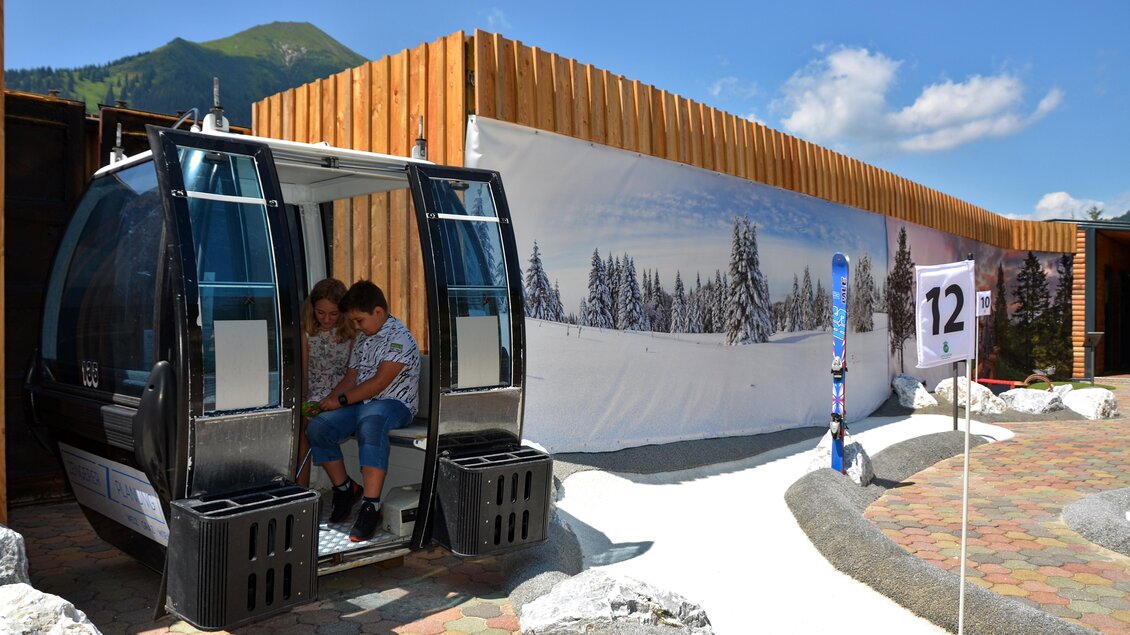 Eine Skiliftgondel steht bereit, während zwei Kinder darin sitzen. Im Hintergrund sieht man eine verschneite Landschaft und eine Schautafel mit der Nummer 12. | © Mountain Adventure Golf