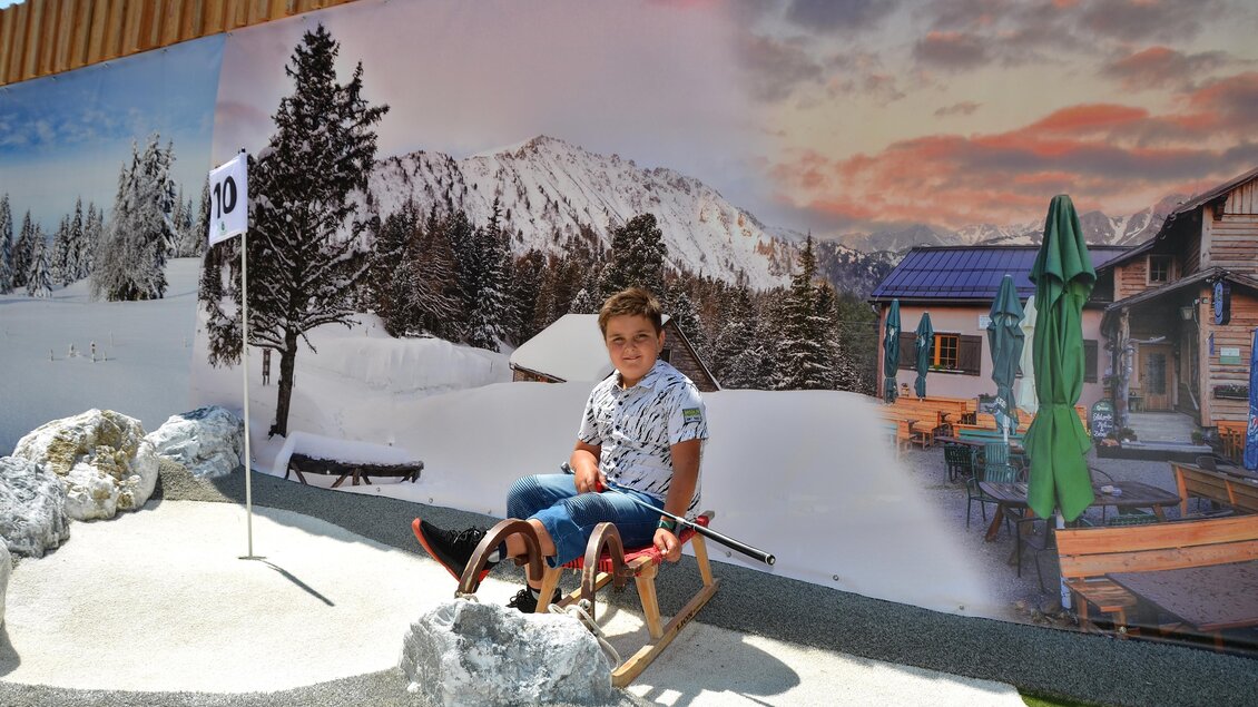 Ein Junge sitzt auf einem Holzschlitten vor einer malerischen Winterlandschaft mit Bergen und schneebedeckten Bäumen. Im Hintergrund ist ein gemütliches Chalet und eine grüne Markise zu sehen. | © Mountain Adventure Golf