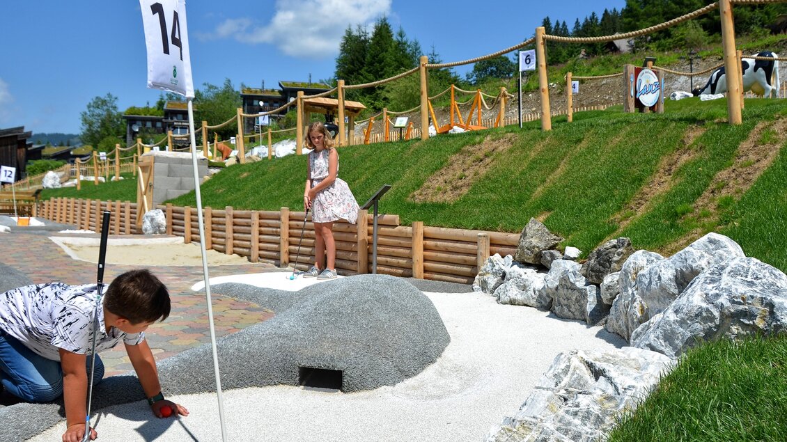 Ein kinderfreundlicher Mini-Golfplatz in einer sonnigen Umgebung. Zwei Kinder spielen und erkunden die Anlage mit vielen grünen Wiesen und Natursteinen. | © Mountain Adventure Golf